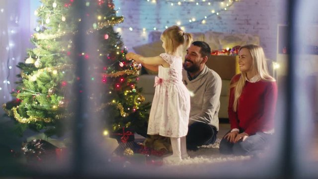 Looking Through Snowy Window. Happy Family: Father, Mother and Their Little Daughter are Decorating Christmas Tree. Shot on RED Cinema Camera 4K (UHD).