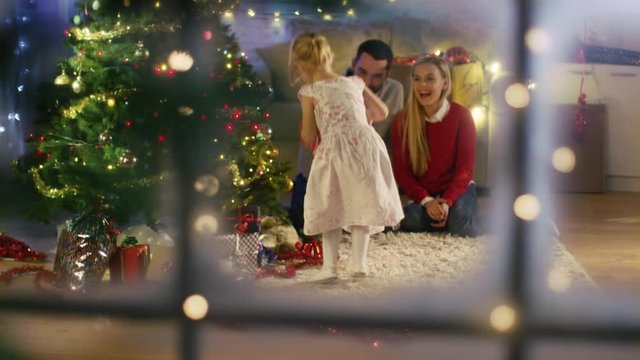 Looking Through Snowy Window. Happy Father, Mother and Daughter Sitting Under Christmas Tree. Daughter Gives a Gift to Her Mother. Shot on RED Cinema Camera 4K (UHD).