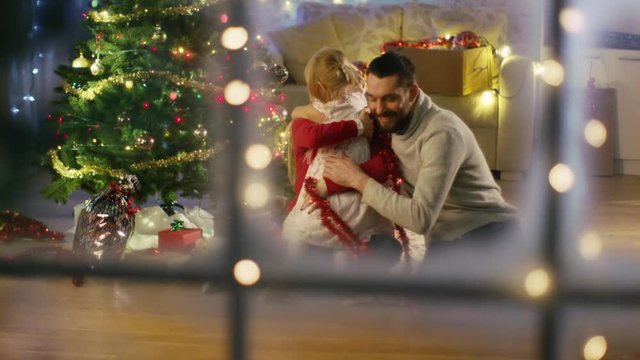 Looking Through Snowy Window. Happy Father, Mother and Daughter Sitting Under Christmas Tree. They All Hug. Shot on RED Cinema Camera 4K (UHD).