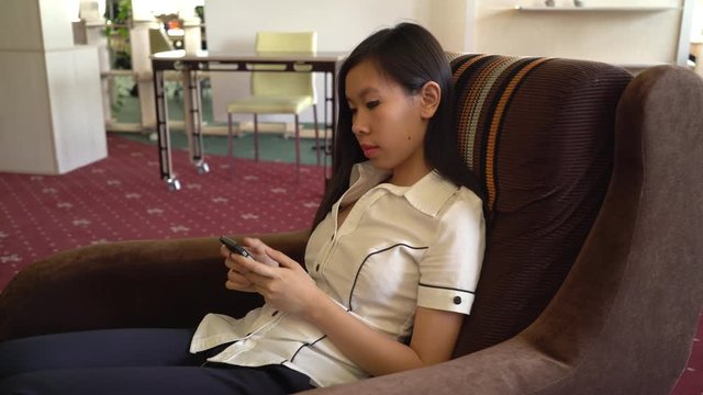 Asian woman texting on smartphone sitting in the chair. Girl in casual dress chatting in social net use cell.