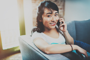 Young happy Asian woman spending rest time at home on sofa and using smartphone for making call.Blurred background, flares effect.