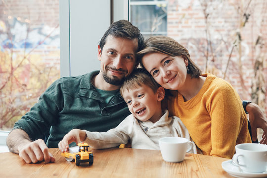 Portrait Of White Caucasian Happy Family Of Three Mother, Father And Son, Sitting In Restaurant Cafe At Table, Smiling Playing, Authentic Lifestyle