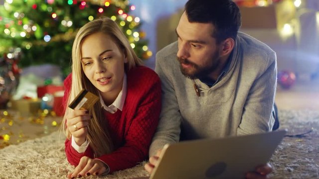 Happy Couple Lying On the Carpet Under Christmas Tree, Woman Holds Credit Card and Man Buys Her Presents on the Laptop. Shot on RED Cinema Camera 4K (UHD).