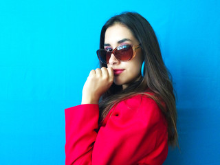 Fototapeta premium Fashion sexy model in sunglasses, beautiful young woman with hand in her mouth with red coat clothes and long brunette hair in a bright vibrant blue color