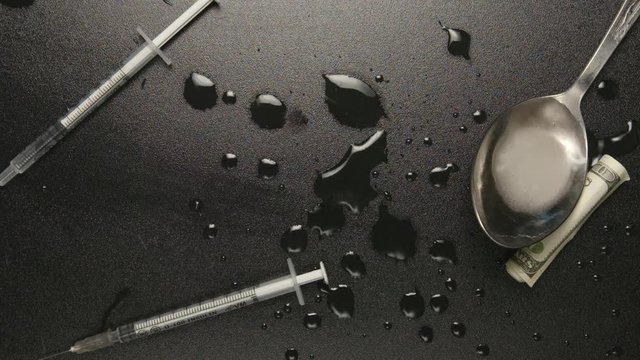 TOP VIEW: Spoon with heroin falls on a black table with syringes (slow motion)
