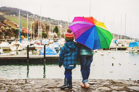 Two cute kids resting by the lake on a rainy day, hiding under big colorful umbrella, back view. Image taken in Lutry, canton of Vaud, Switzerland