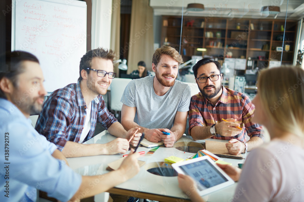 Wall mural Group of young men sitting close together at table during meeting in modern spacious office, listening intently to their female boss, suggesting ideas and discussing data