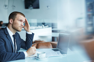 Handsome young businessman wearing formal attire working during coffee break in modern restaurant hall:  looking up from documents with tired and thoughtful expression, rubbing his forehead