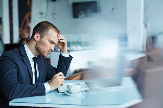 Young handsome man wearing formal attire working with laptop in empty hall of modern restaurant during coffee break, looking puzzled and thinking hard trying to solve some important matter