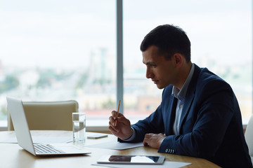 Profile view of young business man sitting against window at table in office, working with laptop and making notes on paper looking focused