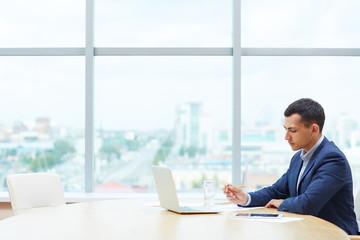 Profile view of young business man sitting alone against window at large table in office, revising...