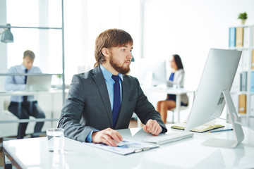 Portrait of bearded  businessman looking at computer screen while sitting at workplace in modern office with other people blurred in background