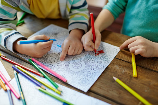 Close-up shot of two pairs of little hands coloring mandala with felt-tip pens, pencils of different colors and illustrations laid on wooden table