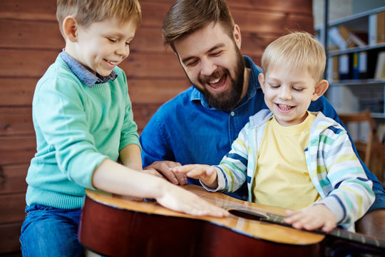 Young bearded father having fun with his sons: little boys with smiles on their faces tapping rhythm on acoustic guitar while their parent looking at them joyfully