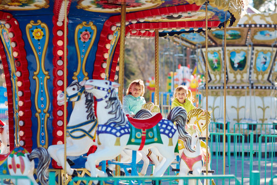 Portrait of funny fair-haired little girls with wide smiles riding on colorful carousel while spending weekend with their family in park