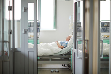 Wide shot image of doors opened to sterile light hospital ward with sad lonely old woman lying on...