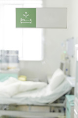 Image of empty bed in clinic with white clean sheets tossed over as if patient just got up and left, shot through glass door with green Hospital Ward sticker on it