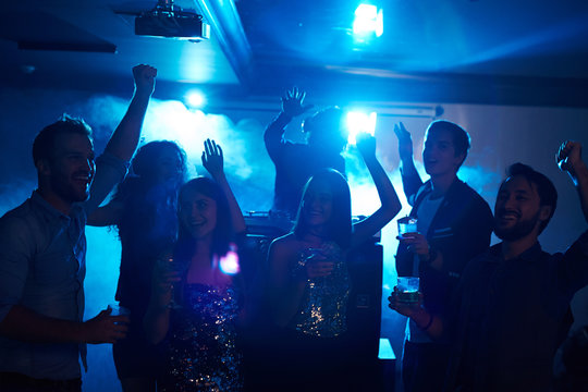 Happy Students Celebrating Graduation In Night Club, Dancing In Club Smoke With Alcohol Cocktails Beside DJ Booth And Having Fun