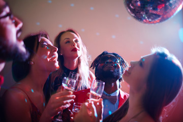 Head and shoulders portrait of three young women and their two male friends holding champagne flutes in hands and looking at disco ball with delight