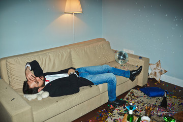 Asian man in jeans and jacket having nap in messy living room after wild New Year party