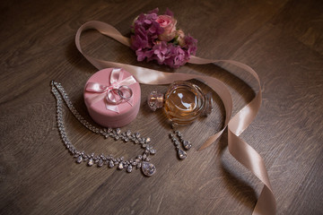 Wedding accessories for bride: wedding rings on pink box, perfume bottle, jewelry and flowers with peach ribbon on wooden background with copy space