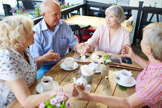 Cheerful senior people sitting in outdoor cafe and holding hands, worn-out photo book lying on table