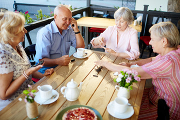 Smiling elderly people having tea party in lovely outdoor cafe and playing dominoes enthusiastically
