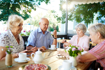 Senior friends gathered together in summer cafe in order to drink tea with pastry and playing dominoes