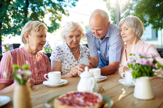 Three senior women and one senior man spending time in outdoors cafe with tea and pastry and watching photos on smartphone - Powered by Adobe