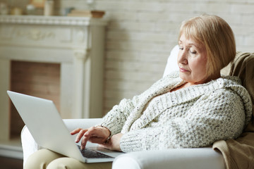 Portrait of modern retired woman in warm home clothing sitting by fireplace with laptop on her laps