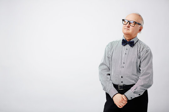 Portrait Of Stylish Elderly Man Standing With His Hands Clasped And Looking Upwards Pensively Against White Background