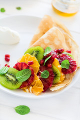 Thin crepes with fresh kiwi, orange, sicilian orange, honey, cream and mint leaves on white rustic wooden table. Selective focus