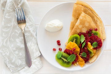 Thin crepes with fresh kiwi, orange, sicilian orange, honey, cream and mint leaves on white rustic wooden table. Selective focus