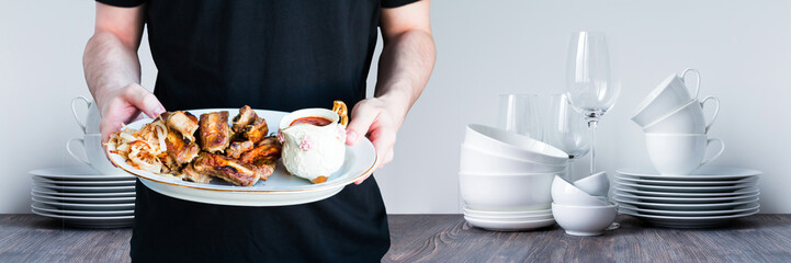 Young man with barbecued spareribs, pork ribs, tomatoe sauce and roast onion slices on white porcelain dish. Restaurant kitchen white background. Selective focus.