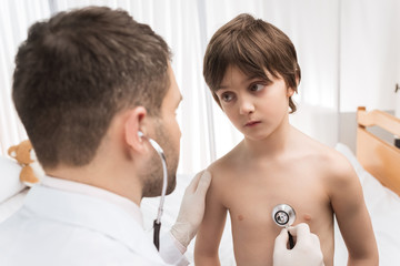 Man doctor examining little boy with stethoscope in hospital