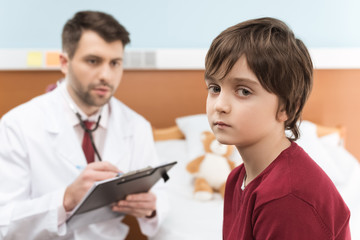 Bored little boy sitting near man doctor with stethoscope and clipboard
