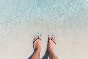 Male feet wearing female flip flops at beach - obrazy, fototapety, plakaty