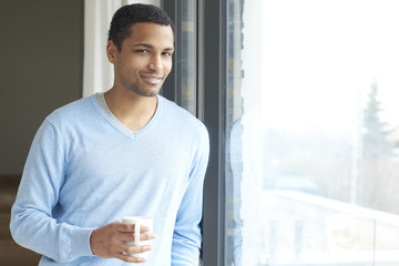 Confident young man portrait. Shot of a young man relaxing by the window at his modern home with a cup of tea.  