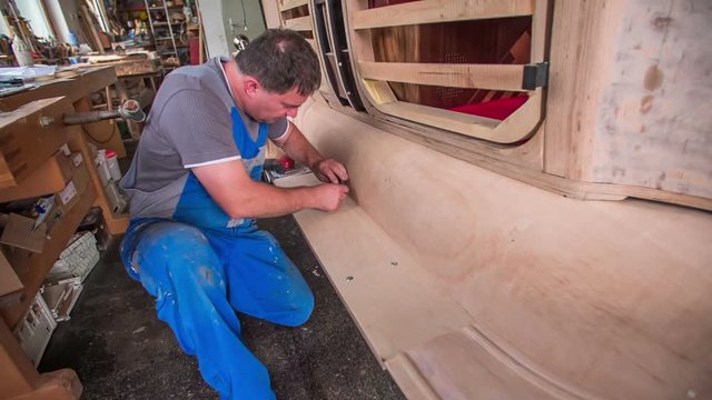 A joiner is sitting down. He is making sure that the surface of the wooden car is flat and polished. He is using a steel wool.
