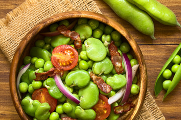 Broad bean, green pea, cherry tomato, red onion and fried bacon salad in wooden bowl, photographed overhead on dark wood with natural light