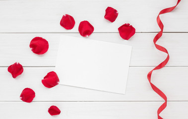 Rose petals, red ribbon and empty paper on white wooden background. Flat lay, top view.