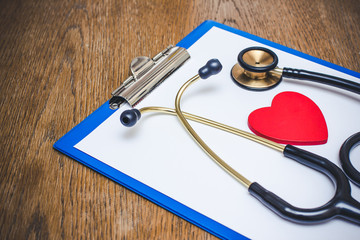 Doctor clipboard stethoscope and heart shape on white paper. Brown wooden table.