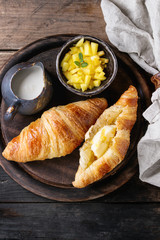 Breakfast with two croissant, butter, cream and sliced mango fruit, served on wood round serving board with textile napkin over old wooden background. Top view with space.