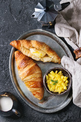 Breakfast with two croissant, butter, coffee maker, cream and sliced mango fruit, served on serving metal tray with textile napkin over black concrete texture background. Top view with space.
