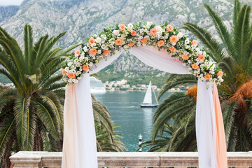 Obraz na płótnie Canvas Wedding ceremony with flower arch near palm, sea, yacht, mountains. Flower decoration is from cream, orange, white roses.