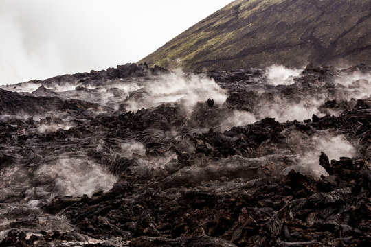 Barren lava fields steaming in light rain with green volcano slope in background