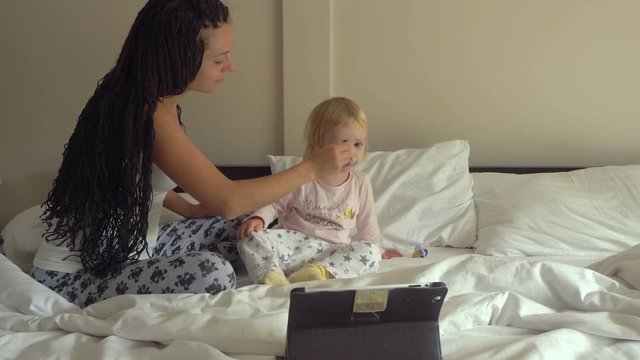 Young pretty woman feeding little baby girl porridge while looking on tablet computer