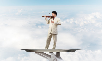Businessman on metal tray playing violin against blue sky background
