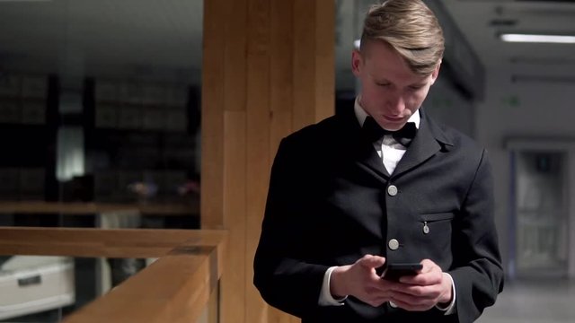 Young attractive businessman standing near the stairs and uses a smart phone. He is dressed in an expensive suit with a bow tie and looks very successful. Waiting for someone