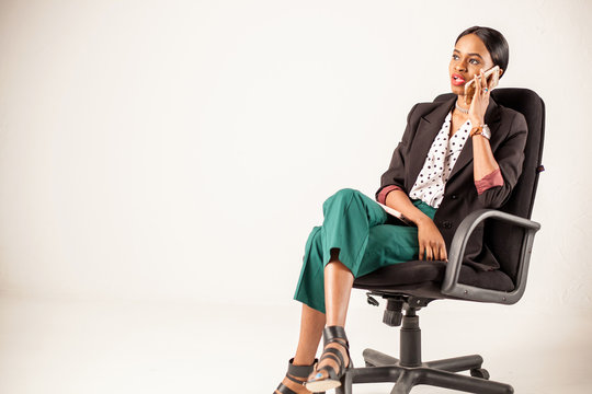 Afroamerican Woman In Business Clothing Sitting In The Chair On A White Background And Talking On The Phone.